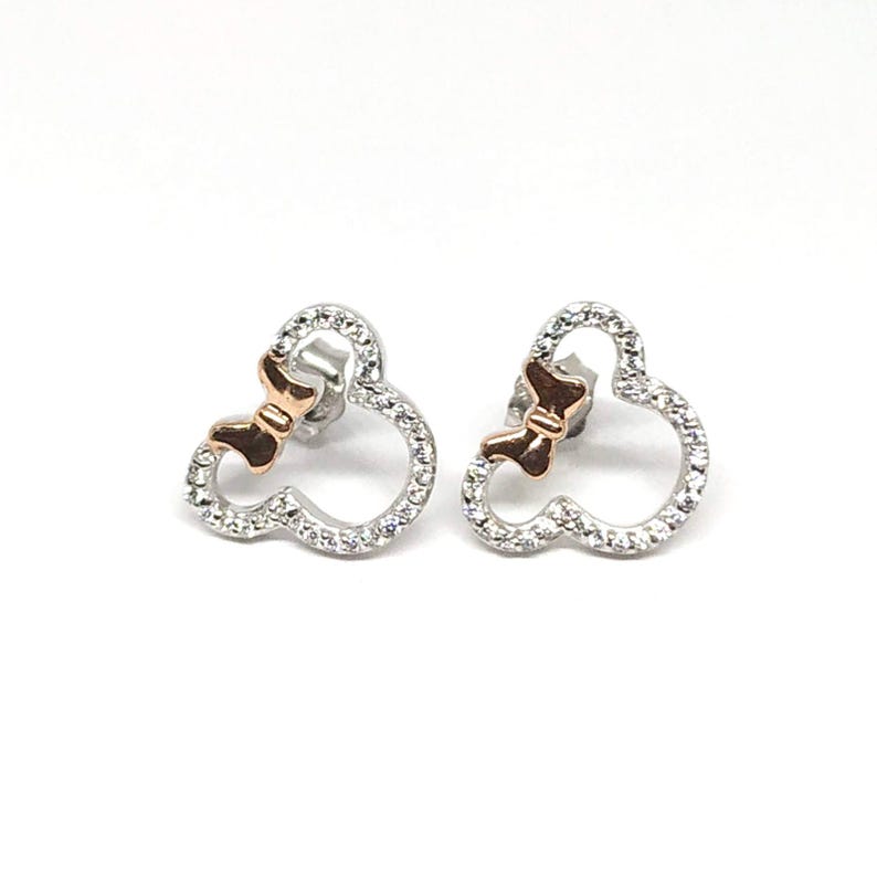 May include: A pair of silver stud earrings. Each earring is shaped like a mouse head with a bow. The mouse head outline is encrusted with small, clear crystals. The bow is a rose gold colour. The earrings are on a white background.