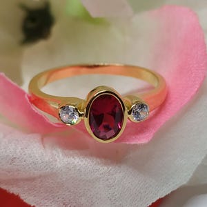 Op de afbeelding: Een gouden ring met een grote ovale robijn en twee kleinere ronde witte edelstenen aan weerszijden.