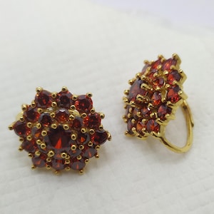 Op de afbeelding: Een paar gouden oorbellen met rode edelsteen accenten. De oorbellen hebben de vorm van een bloem met meerdere lagen edelstenen.