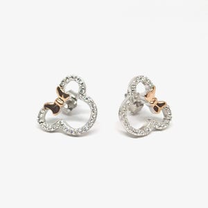 May include: A pair of stud earrings in the shape of Mickey Mouse. The earrings are encrusted with small, clear crystals and feature a rose gold bow. The earrings are displayed on a white background.