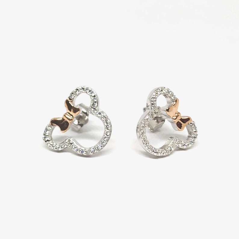 May include: A pair of stud earrings in the shape of Mickey Mouse. The earrings are encrusted with small, clear crystals and feature a rose gold bow. The earrings are displayed on a white background.