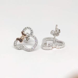 May include: A pair of stud earrings shaped like a mouse head with a bow. The earrings are silver-toned with a gold-toned bow and are encrusted with small, clear crystals. The earrings are presented against a white backdrop.