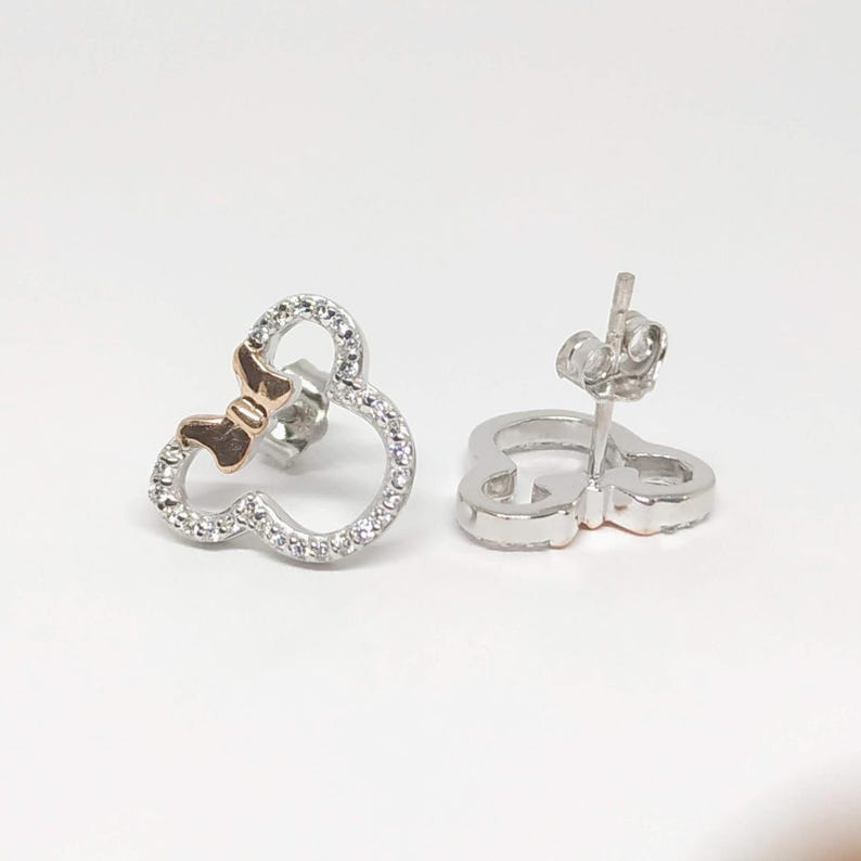 May include: A pair of stud earrings shaped like a mouse head with a bow. The earrings are silver-toned with a gold-toned bow and are encrusted with small, clear crystals. The earrings are presented against a white backdrop.