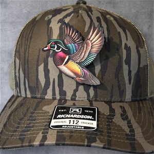 May include: A camouflage trucker hat with a colorful wood duck emblem. The hat features a curved brim, mesh back, and an adjustable strap. The Richardson brand label is visible on the front.