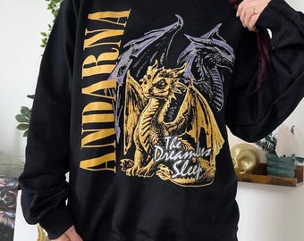 May include: A black long-sleeve sweatshirt featuring a large graphic of a golden dragon with purple-accented wings. The text "ANDARNA" is printed vertically in gold on the left, and the words "The Dreamless Sleep" appear in white script.