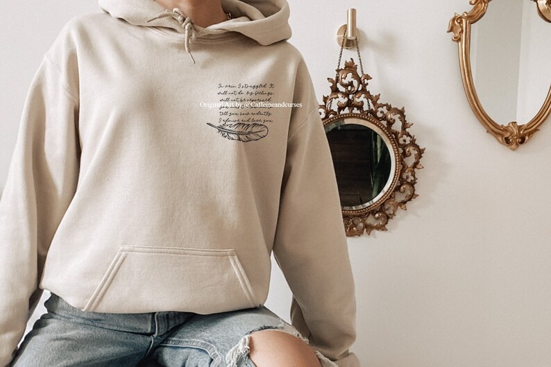 May include: A beige hoodie with a black feather and a quote that reads "In pain I've learned that I shall not be by my feelings, and what I've encountered will not be forgotten. I'll give you my divinity, I'll always love you."