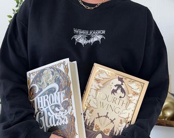 May include: A black crewneck sweatshirt featuring white embroidered text that reads WINGLEADER above a detailed illustration of dragon wings. The sweatshirt is worn with blue denim jeans and is paired with two hardcover books titled Throne of Glass.