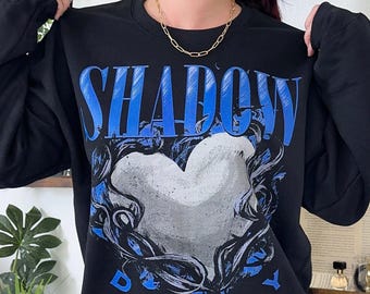 May include: A black long-sleeve sweatshirt featuring the word SHADOW in large blue letters at the top, a central graphic of a white heart surrounded by dark swirling lines, and the word DADDY in blue letters underneath the heart graphic.