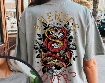 May include: A grey short-sleeve t-shirt featuring a graphic print of two intertwined snakes, one red and one yellow, surrounded by white flowers and black leaves. The text "ENEMIES" appears in yellow above and "LOVERS" in red below the graphic.