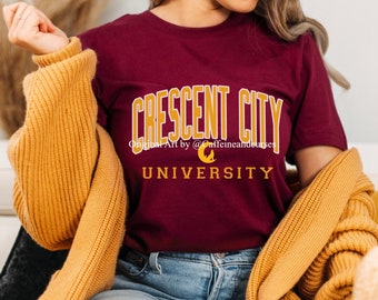 Puede incluir: Una camiseta color burdeos con un gráfico dorado que dice "Crescent City University" y "Original Art by @Caffeineandurses".