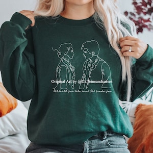 Puede incluir: Una sudadera verde oscuro con un dibujo de línea blanca de una pareja. El texto "Original Art by @Caffeineandcurses" y "She loved you ever since she knew you" está impreso en la sudadera.