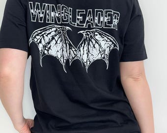 May include: A black short-sleeved t-shirt featuring a white graphic print on the back. The design includes the text "WINGLEADER?" in a bold, outlined font positioned above a detailed illustration of two spread bat-like wings with textured shading.