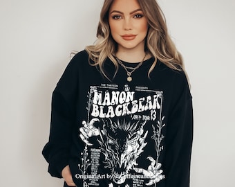 May include: Black sweatshirt with a white graphic print of a concert poster for "Manon Blackbear World Tour 13". The poster features a black and white illustration of a dragon with a menacing expression. The text "The Thirteen Presents" is at the top of the poster.