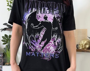May include: A black short-sleeved t-shirt featuring a graphic print of a winged figure in a dynamic pose. The text WINGSPAN is printed in white above the figure, and the word MATTERS is printed in purple below, set against a purple and white cloud background.