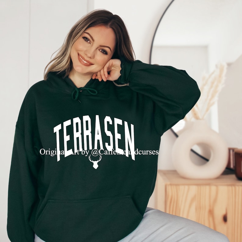 Puede incluir: Una sudadera con capucha verde oscuro con la palabra "TERRASEN" en texto blanco en la parte delantera. El texto est&aacute; en estilo arqueado. Debajo del texto hay un gr&aacute;fico de cabeza de ciervo blanco. El texto "Original art by @Caffeineandcurses" est&aacute; impreso debajo del gr&aacute;fico.