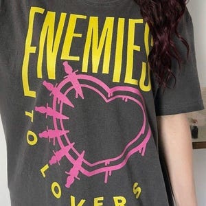 Puede incluir: Una camiseta gris carbón de manga corta con el texto amarillo ENEMIES TO LOVERS y un gráfico de corazón rosa delineado con pequeñas ilustraciones de dagas. El diseño está centrado en la parte frontal de la camiseta con un estilo gráfico.