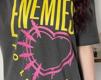 May include: A charcoal grey short-sleeve t-shirt featuring the yellow text ENEMIES TO LOVERS and a pink heart graphic outlined with small dagger illustrations. The design is centred on the front of the shirt with a bold, graphic style.