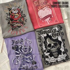 Puede incluir: Cuatro dise&ntilde;os diferentes de camisetas con estampados gr&aacute;ficos. La primera camiseta es gris con una serpiente roja y flores y el texto "Enemies Lovers". La segunda camiseta es roja con un coraz&oacute;n y llamas y el texto "Slow Burn Romance". La tercera camiseta es morada con una mano y el texto "Fated Mates". La cuarta camiseta es gris con una calavera y el texto "Morally Grey World Tour Who Did This To You Touch Her And Die".