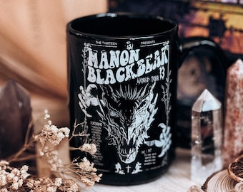 May include: A black ceramic mug featuring a white graphic print of a dragon head and the text "The Thirteen Presents Manon Blackbeak World Tour 13" along with a list of names and a small cross symbol centred at the bottom of the design.
