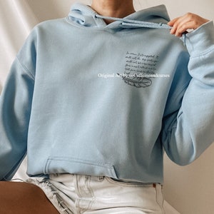 May include: Light blue hooded sweatshirt with a black feather and text that reads "So when I struggled it still was to be bigger than what the world told me I should be.  The you you were made for was larger than what you see."  Original Art by @Caffeineandcurses