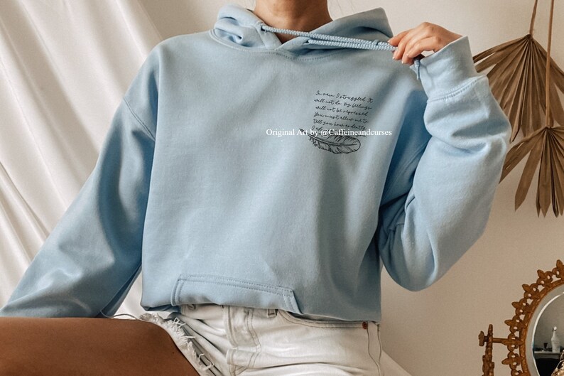 May include: Light blue hooded sweatshirt with a black feather and text that reads "So when I struggled it still was to be bigger than what the world told me I should be.  The you you were made for was larger than what you see."  Original Art by @Caffeineandcurses