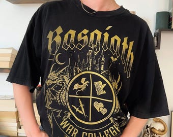May include: A black short-sleeved t-shirt featuring a gold graphic print. The design includes the text "Basgiath War College" in a gothic font, a castle silhouette, a dragon, and a circular emblem containing a dragon, scroll, daggers, and hearts.