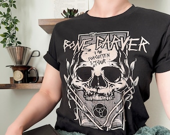 May include: Black t-shirt with a graphic of a skull and the text "The Bone Carver I Am Forgotten Tour" printed on the front.