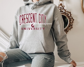Puede incluir: Una sudadera con capucha gris con el texto "Crescent City University" en letras color granate. El logotipo de la universidad es una luna creciente con una estrella en su interior.