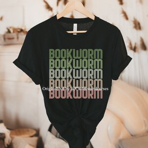 May include: Black t-shirt with a graphic design of the word "BOOKWORM" repeated five times in a retro style with green, yellow, and pink colors. The text "Original Art by Caffeine and Curses" is visible at the bottom of the design.