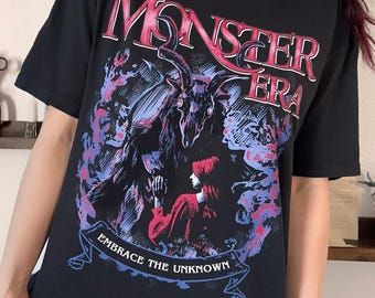May include: A black short-sleeved t-shirt featuring a graphic print with the text MONSTER ERA at the top and EMBRACE THE UNKNOWN on a banner at the bottom. The illustration depicts a horned creature and a figure in red surrounded by purple smoke.