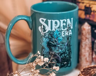 May include: A teal ceramic mug featuring a white graphic print that reads "SIREN ERA" above an illustration of a mermaid surrounded by seaweed and bubbles. The mug has a curved handle and a smooth finish, showcasing a detailed aquatic design.