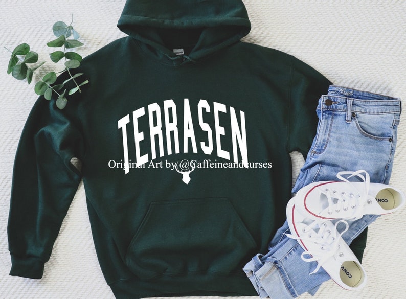 Puede incluir: Una sudadera con capucha verde oscuro con la palabra "TERRASEN" en texto blanco en la parte delantera. El texto est&aacute; en una fuente arqueada. Debajo del texto est&aacute; la frase "Original Art by @caffeinandcurses" en texto blanco. Un gr&aacute;fico de cabeza de ciervo blanco est&aacute; debajo de la frase.