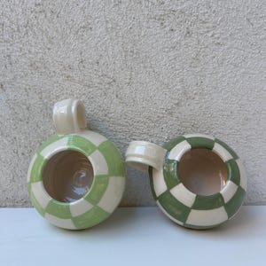 May include: Two ceramic mugs with a round shape and a handle. The mugs are painted with a checkerboard pattern in light green and cream colors. The interior of the mugs is a light beige color.