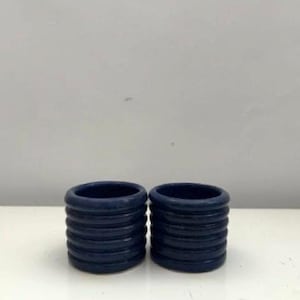 May include: Two cylindrical napkin rings in a deep navy blue colour. Each ring features a series of five horizontal, raised circular ridges that wrap around the exterior, creating a textured, ribbed appearance with a smooth, matte finish.