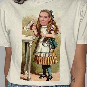 May include: Cream-colored t-shirt featuring a vintage illustration of a woman dressed as Alice in Wonderland, holding a sign that reads "DEUTSCHLAND". The artwork is in a classic storybook style, with a whimsical design and a touch of humor.