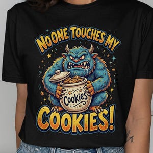 May include: A black short-sleeve t-shirt featuring a graphic of a blue, furry, horned monster holding a cookie jar. The text above reads NOONE TOUCHES MY and the text below reads COOKIES! with the word Cookies printed on the jar itself.