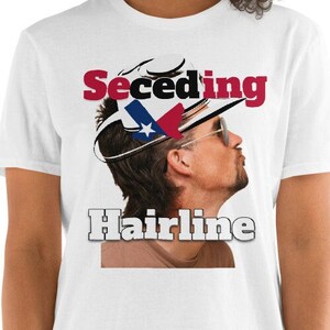Puede incluir: Camiseta blanca con un gráfico rojo y azul de un hombre con una línea de cabello que retrocede usando un sombrero de vaquero con la bandera de Texas en él. El texto "Seceding Hairline" está impreso en la camiseta.