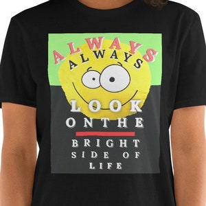 Op de afbeelding: Zwart T-shirt met een grafische print van een gele smiley met de tekst "ALWAYS LOOK ON THE BRIGHT SIDE OF LIFE" in rood, groen en wit.
