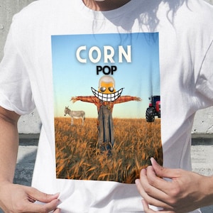 May include: White t-shirt with a graphic of a scarecrow in a field of wheat. The scarecrow has a smiling face and the text "CORN POP" above it.