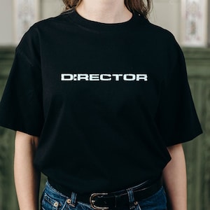 Può includere: T-shirt nero con la scritta "DIRECTOR" stampata in bianco sul petto. La maglietta ha maniche corte e girocollo. La persona indossa jeans blu e una cintura nera.