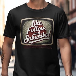 Op de afbeelding: Zwart T-shirt met een vintage televisiescherm afbeelding met de tekst "Like, Follow, and Subscribe!"