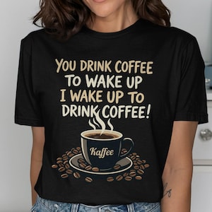 Peut inclure: T-shirt noir avec l'inscription "You drink coffee to wake up, I wake up to drink coffee !" et un motif de tasse à café. La tasse est bleu foncé avec le mot "Kaffee" dessus, entourée de grains de café.