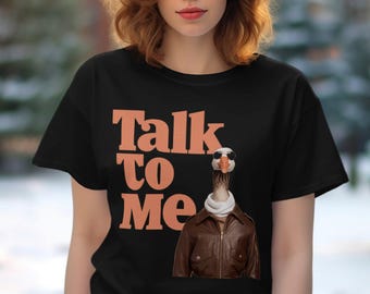 以下が含まれることがあります： オレンジ色の文字で「Talk to Me」と書かれた黒いTシャツ。サングラスと茶色の革のジャケットを着たガチョウがテキストの前に立っています。
