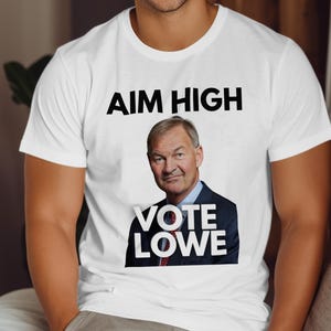May include: White t-shirt with the text "AIM HIGH" above a portrait of a man in a suit. Below the portrait, the text reads "VOTE LOWE". The shirt is a crew neck style.