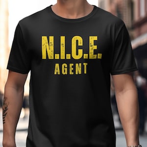 以下が含まれることがあります： 黒いTシャツ。黄色の文字で「N.I.C.E. AGENT」と書かれています。Tシャツはクルーネックです。背景はぼやけた屋外のシーンです。