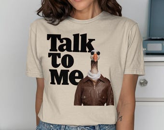 以下が含まれることがあります： ベージュのTシャツに黒い文字で「Talk to Me」と書いてあります。テキストの下には、パイロットサングラスと茶色の革のジャケットを着たガチョウが描かれています。
