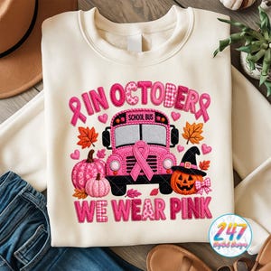 May include: A cream-colored crewneck sweatshirt featuring a graphic design with a pink school bus, pumpkins, autumn leaves, and hearts. The text reads "IN OCTOBER WE WEAR PINK" with pink awareness ribbons integrated into the design elements.