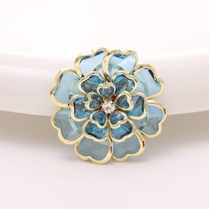 Puede incluir: Broche con forma de flor con pétalos azul claro y turquesa, contorneados en dorado. El centro de la flor presenta un pequeño cristal brillante. Accesorio decorativo.