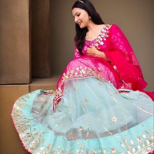 May include: A traditional Indian outfit featuring a pink top and a light blue skirt, both adorned with gold embellishments. The outfit includes a bright pink scarf. The ensemble is worn by a person.