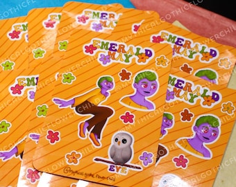 May include: Set of five stickers featuring a cartoon character with purple skin and green hair. The character is wearing a yellow shirt and brown pants. The stickers are on a yellow background with orange stripes and green and orange flowers. The text "Emerald May" is printed on the stickers.